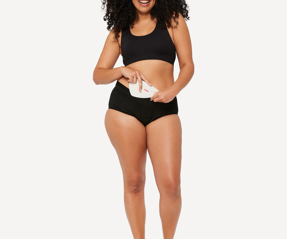 Woman in black Scarlet underwear sliding the white rae heat pad into the pocket of her brief